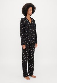 Set de pijama negru cu imprimeu floral, compus dintr-o cămașă cu guler și mâneci lungi, și pantaloni asortați. Materialul pare moale și ușor.