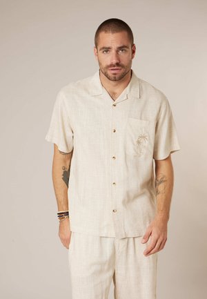 Homme aux cheveux courts et à la barbe, portant une chemise beige à manches courtes boutonnée et un pantalon assorti, debout devant un fond uni.