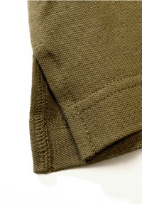 Gros plan d'un tissu tricoté vert olive montrant un ourlet replié avec des coutures visibles et une texture sur fond blanc.