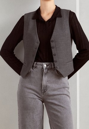 Grau maßgeschneiderte Weste über einem schwarzen Langarmshirt, kombiniert mit hochtaillierten grauen Jeans. Die Weste hat sechs Knöpfe und eine strukturierte Passform.