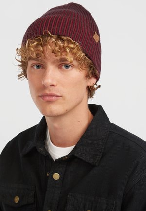 Gebreide muts in bordeauxrood met een gestructureerd verticaal streeppatroon. Heeft een klein, lichtbruin label. Model draagt een zwart ribfluwelen jasje.