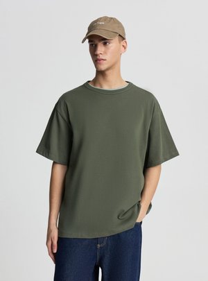 Giovane uomo che indossa una t-shirt oversize verde oliva, jeans blu scuro e un cappellino da baseball beige con la scritta "Las Vegas", in piedi con una mano in tasca.