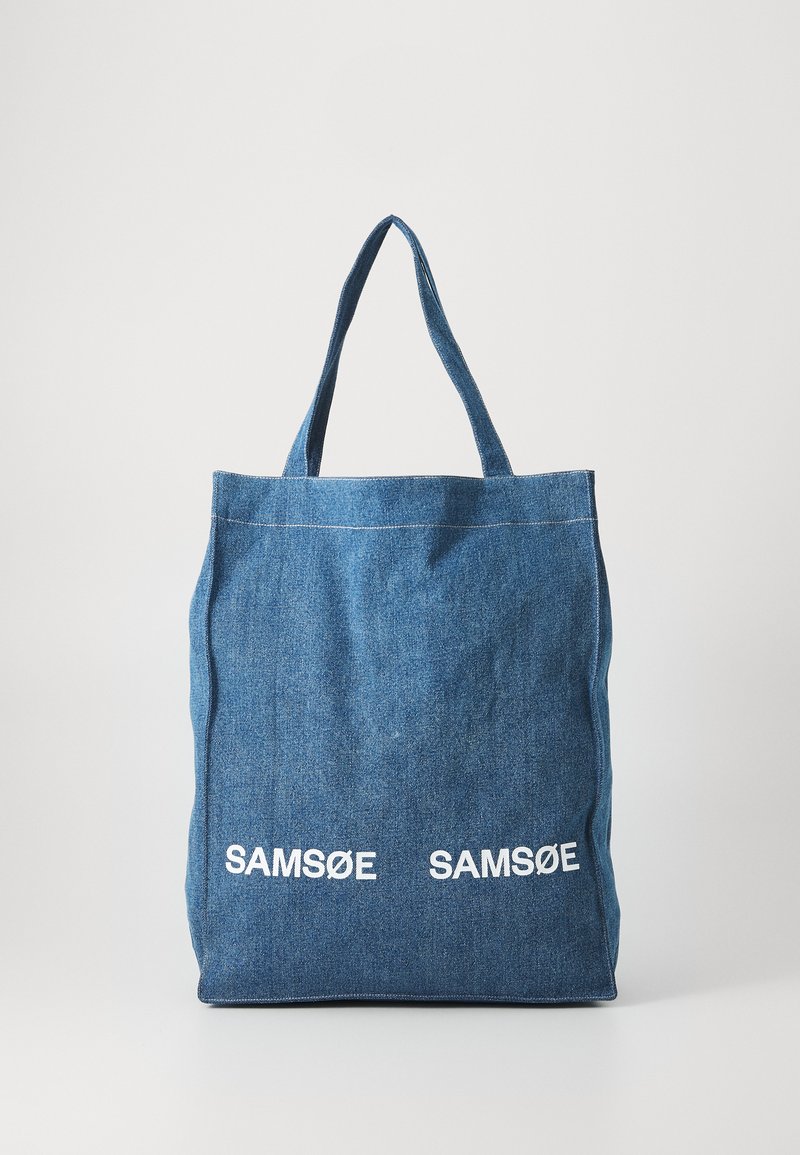 Tamsiai mėlynas džinsinis tote maišas su dviem vertikaliais baltos spalvos "SAM SøE" tekstiniais logotipais. Yra plačios rankenos ir paprastas, nesuformuotas dizainas.