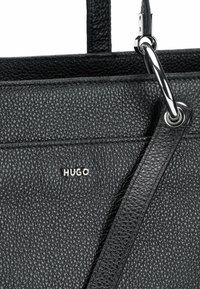 Borsa a mano in pelle nera con superficie strutturata, caratterizzata da un logo metallico argentato "HUGO" e una tracolla in pelle staccabile con dettagli in metallo argentato.