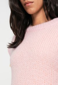 Femme aux longs cheveux foncés portant un pull en maille doux rose pâle, montrée des épaules aux lèvres sur un fond neutre.