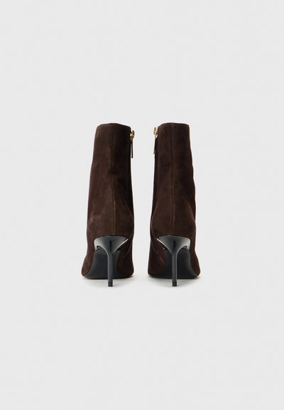 Hnedé semišové ankle boots so zoštíhľujúcim tvarom, vysokými stiletto opätkami a zlatými zipsami na zadnej strane pre uzatvorenie a detail.