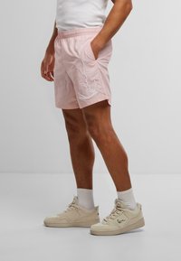 Rosa bomullsshorts med elastisk midje, sidelommer og hvit outline-design på den ene siden, kombinert med beige joggesko og hvite sokker.