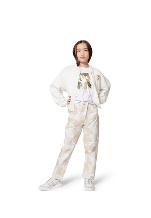 Giacca bomber bianca su una camicia bianca con stampa dorata, abbinata a pantaloni beige con fantasia e sneakers bianche. L'outfit presenta design e texture moderne.