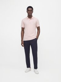 Hombre con una camisa tipo polo rosa claro de manga corta, pantalones azul marino oscuro y zapatillas blancas, de pie con una mano en el bolsillo.