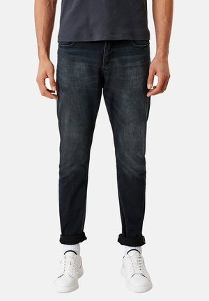 Jeans de mezclilla azul oscuro con corte ajustado. Presentan un ligero desgaste, diseño clásico de cinco bolsillos y puños enrollados. Combinados con zapatillas blancas.