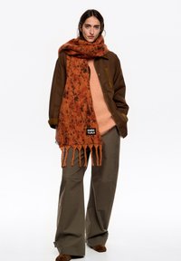 Pomarańczowo-brązowy, nakrapiany szal z wełny z frędzlami na końcach, noszony na beżowym swetrze i brązowej kurtce. Look dopełniają oliwkowozielone, szerokie spodnie.