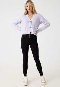 Cárdigan corto de lavanda claro con textura acanalada, cierre de botón y bolsillos frontales, combinado con leggings negros y zapatillas blancas.