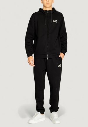 Felpa nera con zip e pantaloni coordinati realizzati in tessuto morbido, con logo EA7 bianco sul petto e sul lato. Sneakers bianche.