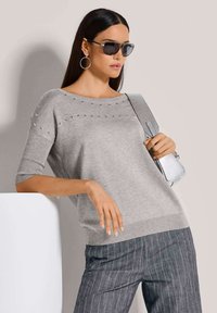 Grauer Pullover mit kurzen Ärmeln, verziert mit metallischen Nieten am Ausschnitt. Kombiniert mit gestreiften Hosen und einer silbernen Handtasche.