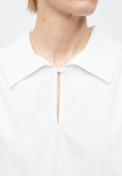 Közeli kép egy fehér inges személyről, melynek széles gallérja és kis gombbal záródó kulcslyuk alakú nyakkivágása van.