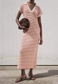 Femme portant une robe midi rayée crème et rouge et des sandales à talons marron, tenant un sac à main tressé marron foncé, debout en plein air.