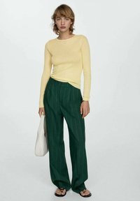 Rumena rebrasta majica dolgih rokavov v kombinaciji s širokimi hlačami z zelenimi črtami. Model drži belo torbico in nosi črne sandale na ravnem.