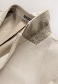 Col de veste beige avec un tissu doux, ressemblant à de la laine, renforçant le bord intérieur sur un fond neutre.