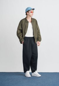 Olivzaļš bombere jaka, balts tops bez piedurknēm, melnas sporta bikses un baltas sporta kurpes. Zils cepure un krāsainas saulesbrilles papildina izskatu.