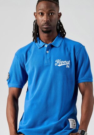 Blauwe polo t-shirt van katoen met korte mouwen. Bevat een witte geborduurde logo "Kaporal 04" op de borst en een badge op de mouw.