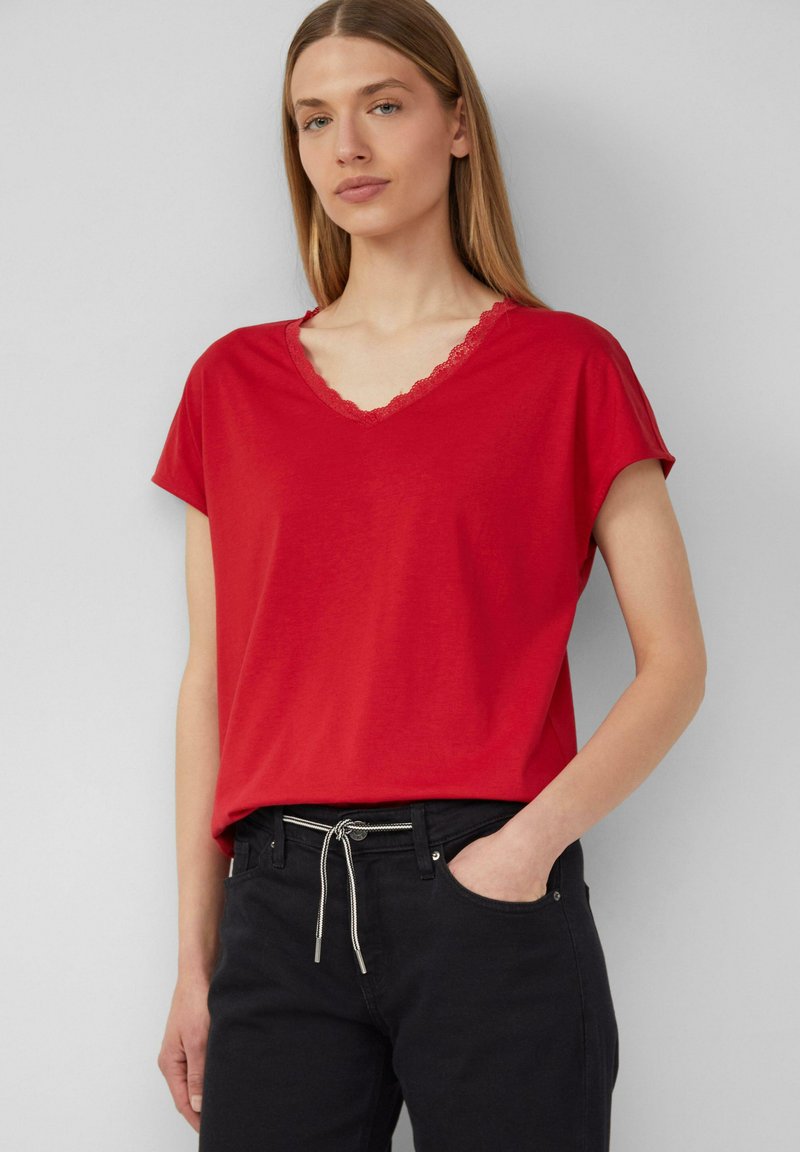 T-shirt rossa a maniche corte con scollo a V e rifiniture in pizzo. Realizzata in un materiale morbido, abbinata a pantaloni neri e una cintura decorativa con cordoncino.