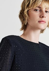 Navy-blaues, strukturiertes Kleid mit Rundhalsausschnitt, das mit schwarzen und silbernen Nieten im gesamten Stoff verziert ist.