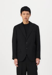 Schwarzer maßgeschneiderter Blazer mit einer glatten Textur, ausgestattet mit einer Zweiknopfverschluss, Klappentaschen und eingeklapptem Revers, kombiniert mit einem schwarzen Hemd.