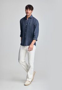 Navy linnen overhemd met lange mouwen, knoopjes aan de voorkant en twee voorkeurs; gecombineerd met witte broeken en lichtbeige espadrilles.