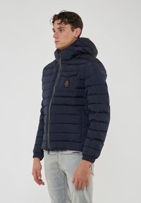 Giacca a vento navy con cappuccio, dotata di segmenti trapuntati, zip frontale, tasca sul petto e marchio sul lato sinistro.
