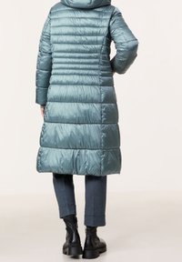 Abrigo acolchado en color azul verdoso con capucha, con un diseño de acolchado vertical, mangas largas y longitud media. Combinado con pantalones oscuros y botas.