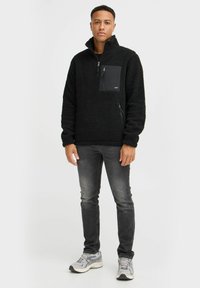 Black fleece pullover with a high collar, front zip pocket, and textured surface, paired with dark gray jeans and athletic shoes.