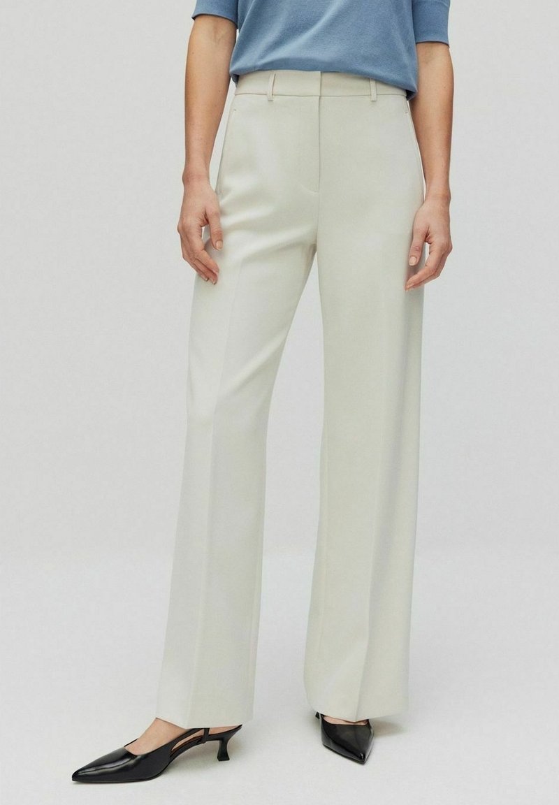 Vidbyxor i ett ljust beige tyg med en slät yta och sidfickor, kombinerade med svarta pumps med spetsig tå.