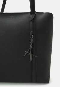 Black leather tote bag with textured surface, featuring elongated handles and a metal logo charm on a chain. Minimalist design.