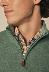 Grüner Strickpullover mit einem Reißverschlusskragen über einem karierten Hemd in Gelb, Marineblau und Weiß. Die strukturierte Stoffbeschaffenheit ist sichtbar.