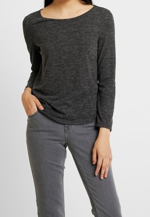 Femme portant un haut à manches longues gris foncé et un jean gris clair, une main dans la poche et l'autre reposant le long du corps.