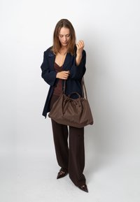 Barna színű, puha anyagból készült zsák, szorosra húzott nyílással és két hosszú fogantyúval, amelyet egy sötétkék blézert és barna ruházatot viselő személy tart.