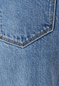 Modrá denimová tkanina s detailním záběrem na kapsu s oranžovými stehy, který zvýrazňuje texturovanou vazbu a ostré úhly designu.