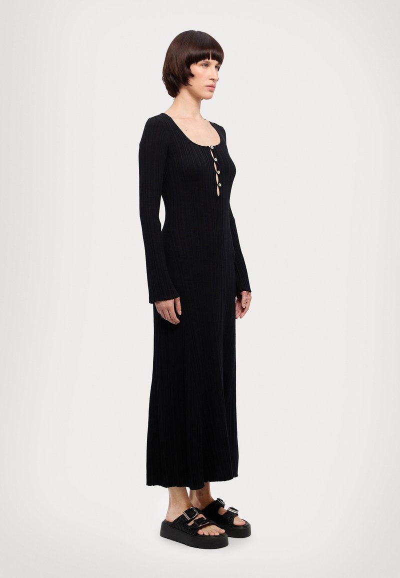Black ribbed long-sleeve dress with a round neckline and button details. The model wears black platform sandals. Smooth texture, floor-length design.