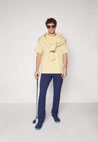 Beige cotton t-shirt, navy trousers, sunglasses, and golf club. Sweater draped over shoulders. Casual athletic outfit.