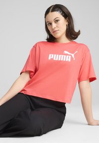 Koralfarvet cropped t-shirt med hvid "PUMA" logo og springende puma grafik. Model iført sorte bukser, glat stof, afslappet pasform.
