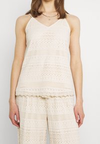 Light beige crochet tank top with spaghetti straps, featuring intricate textured patterns and a scalloped hem, paired with matching pants.