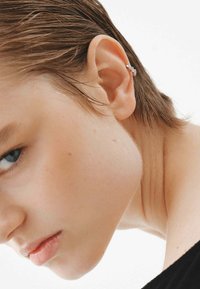 Pendiente de plata en forma de cuff con un diseño en zigzag que presenta pequeños acentos de piedra, exhibido en un primer plano de la oreja y el cuello. Se observa el cabello con textura.