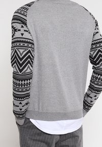 Sudadera gris con mangas negras estampadas con diseños geométricos, cuello y dobladillo acanalados. Material suave con un ajuste relajado.