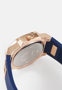 Reloj de oro rosa con correa de silicona azul. Presenta una banda texturizada, caja redonda y una corona a presión en el lateral. Parte trasera plateada con detalles grabados.