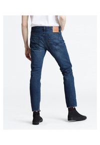 Osoba v slim-fit tmavě modrých džínách a černých vysokých teniskách, stojící zády k fotoaparátu na bílém pozadí.