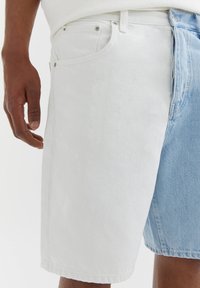 Short en jean avec un design bicolore : un côté gris clair, l'autre bleu clair. Tissu texturé, style cinq poches, coutures visibles.