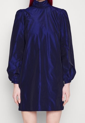 Kobieta ubrana w błyszczącą, ciemnoniebieską, krótką sukienkę z długimi, bufkami i wysokim golfem, stojąca na jednolitym tle.