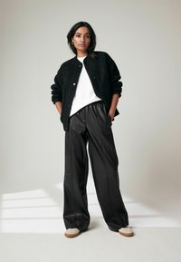 Cardigan noir texturé sur une chemise blanche, associé à un pantalon en cuir noir ample et des chaussures beiges. Design simple avec des détails pratiques.