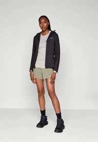 Woman standing wearing a black hooded jacket, light gray t-shirt, olive green shorts, black socks, and black hiking boots on a plain background.