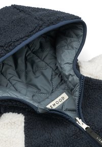 Marineblauwe en witte fleecejas met doorgestikte blauwe binnenvoering, gedeeltelijk open, met het merklabel zichtbaar aan de binnenkant van de kraag.
